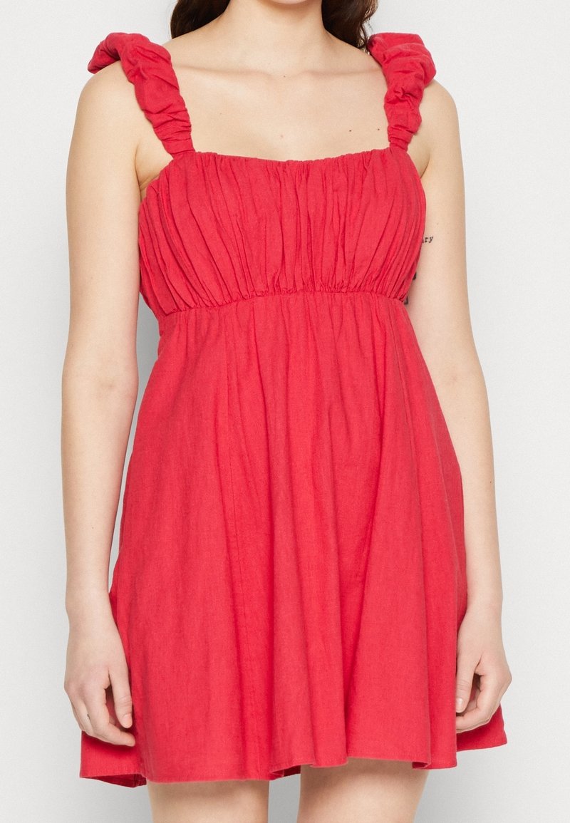 Robe rouge sans manches avec un corsage froncé et des bretelles à volants. Silhouette en A, longueur genou, fabriquée à partir d'un tissu texturé.