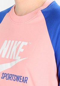 Sportovní tričko Nike s růžovým tělem, modrými raglánovými rukávy, bavlněným materiálem a bílým potiskem loga na přední straně.