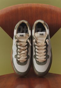 Paire de baskets en daim blanc et vert avec des lacets crème posées sur une chaise en bois avec un fond vert.