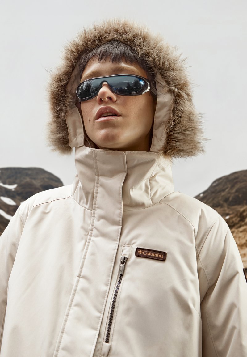 Beige wasserfeste Jacke mit einer gefütterten Kapuze aus Fell, Reißverschluss vorne und Columbia-Logo auf der Brust. Das Model trägt eine Sonnenbrille vor einem Bergpanorama.