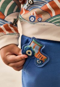 Blaue Baumwollhosen mit einem bunten Traktor-Patch in Orange, Grün und Blau. Die Oberseite hat ein mehrfarbiges Design mit Streifen und Formen.