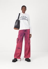 Haut à capuche blanc avec le texte "Original Sportswear", associé à un pantalon cargo rose avec des zips et des jambes larges, complété par des chaussures à plateforme noires.