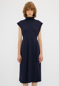 Laivansininen, polvipituinen mekko, jossa on korkea kaula-aukko ja hihattomat olkapäät. Valmistettu pehmeästä kankaasta, jossa on sileä tekstuuri ja liehuva hame.