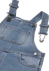 Mono de mezclilla en azul claro con tirantes ajustables, clips de metal y un bolsillo frontal con detalles de costura. Tejido suave y texturizado.