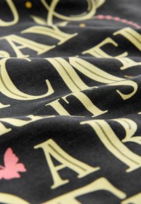 Czarna tkanina z wytłoczonym żółtym napisem i akcentem w postaci motyla w kolorze różowym. Tekstura wydaje się prążkowana, co podkreśla detale nadruku.