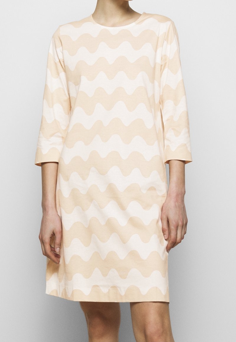 Femme portant une robe beige et blanche à rayures ondulées avec des manches trois-quarts, debout devant un fond clair uni.