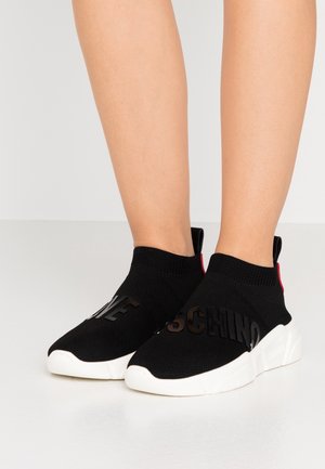 Stopy w czarnych skarpetkowych sneakersach z białymi podeszwami i dużymi wypukłymi literami na wierzchu, stojące na jednolitej jasnej podłodze.