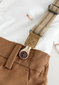 Brown cotton shorts with belt loops and a matching button, paired with a white shirt featuring an embroidered design and beige suspenders.