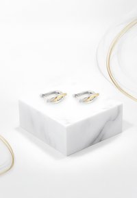 Silvriga manschettknappar med en tvinnad design, med en accent i gult guld och en klar ädelsten, visade på ett vitt marmorpelare.