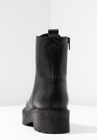 Bottine noire en cuir avec une semelle en caoutchouc texturée, une languette à l'arrière, et une finition lisse. Pas d'ornements ni de logos visibles.