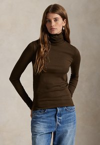 Young woman with long brown hair wearing a dark brown turtleneck and light blue jeans against a plain background.