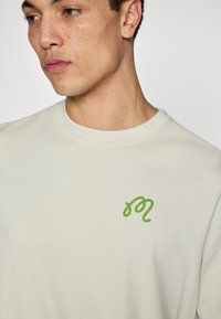 Παλαιά πράσινη βαμβακερή μπλούζα με μικρό πράσινο λογοτύπο με καλλιγραφικά γράμματα στην αριστερή πλευρά του στήθους. Κλασικός στρογγυλός λαιμός και ομαλή υφή.