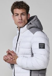 Giacca a vento bianca con dettagli grigi, dotata di colletto alto, cappuccio regolabile, chiusura con zip e patch con logo sulla manica.
