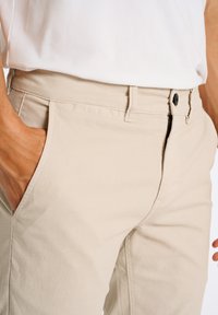 Beige bomuldsbukser med struktureret overflade, der har sidelommer, flad front og en sort knaplukning i livet.