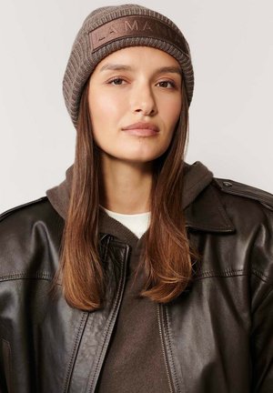 Kobieta z długimi brązowymi włosami w brązowej dzianinowej czapce, brązowej bluzie z kapturem i ciemnobrązowej skórzanej kurtce na jednolitym jasnym tle.