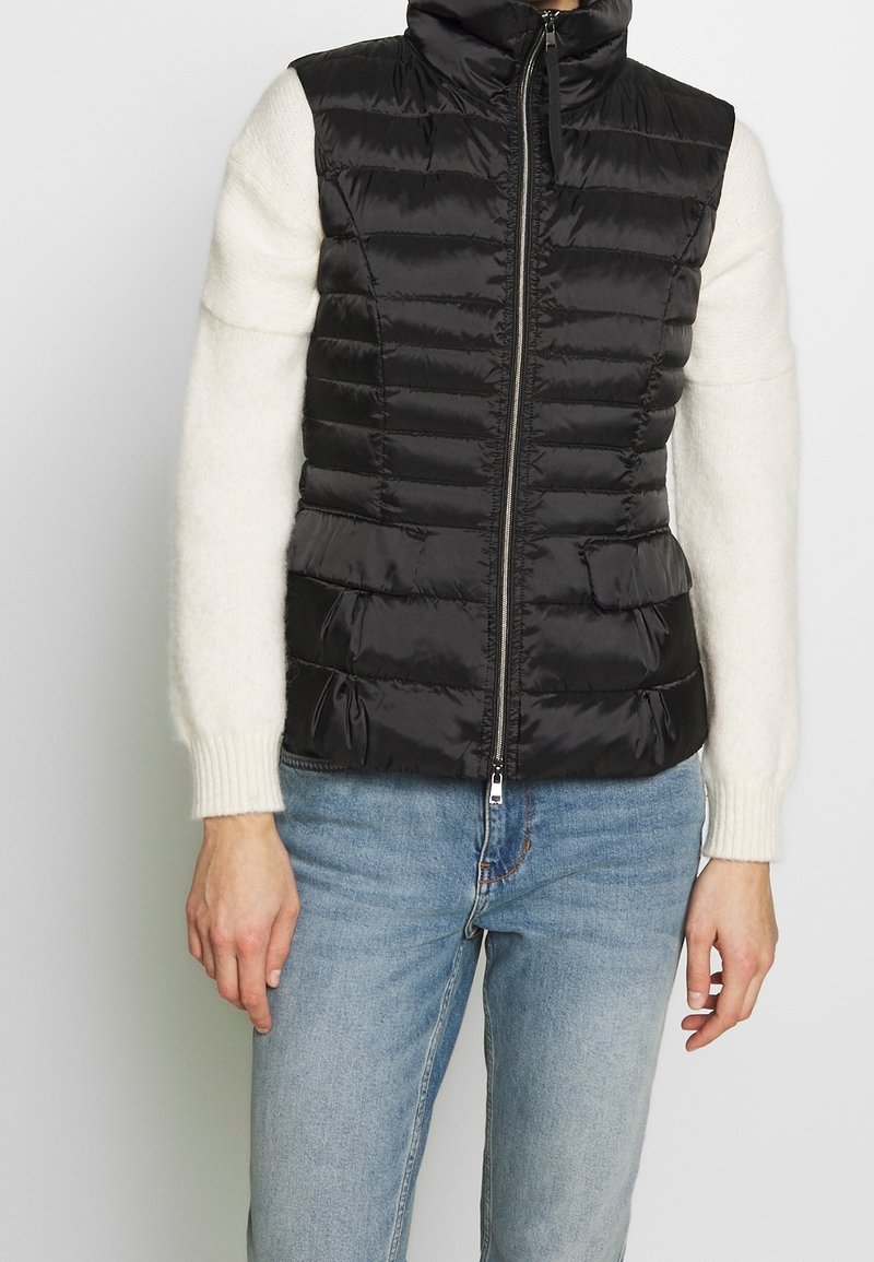 Veste matelassée noire sans manches avec fermeture éclair devant, portée sur un pull blanc à manches longues et un jean bleu.