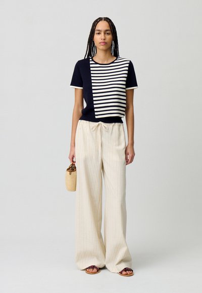 Haut à manches courtes rayé bleu marine et blanc, pantalons larges à rayures beiges, et sandales marron, avec un sac en osier. Coupe décontractée.