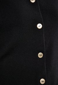 Černý pletený cardigan s řadou pěti knoflíků ve zlatém tónu, hladkým povrchem a přiléhavým střihem.