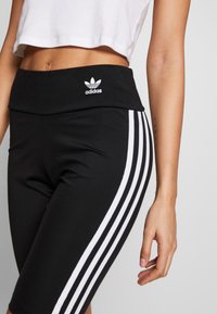 Schwarze Sportleggings mit hohem Bund, drei weißen Streifen an den Seiten und einem gestickten Adidas-Logo am Bund.