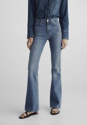 Osoba ubrana w wysokie spodnie dzwony w jasnoniebieskim kolorze, ciemnoniebieską denimową koszulę i czarne sandały ze złotą klamrą na jednolitym szarym tle.