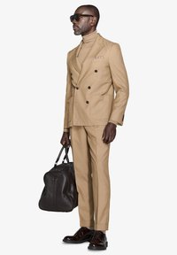 Lys beige dobbeltradet jakkesæt med matchende bukser, turtleneck, brun lædertaske og mørke pæne sko. Glat stof, minimale mønstre.