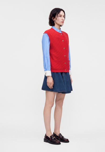 Γυναίκα στέκεται φορώντας κόκκινο αμάνικο γιλέκο με κουμπιά, μπλε πουκάμισο με μακριά μανίκια, τζιν φούστα και σκουρόχρωμα μοκασίνια σε απλό φόντο.