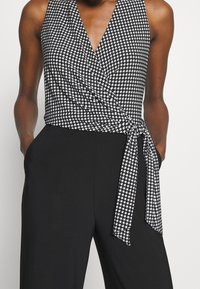 Svartvitt rutig ärmlös topp med v-ringning, med knytning vid midjan och enfärgade svarta byxor. Strukturerad tyg och prydlig finish.