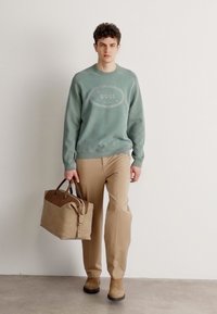 Maglione verde chiaro a coste con logo "BOSS", pantaloni beige e stivaletti alla caviglia in suede marrone. Tiene una borsa in pelle color tan. Sfondo neutro.