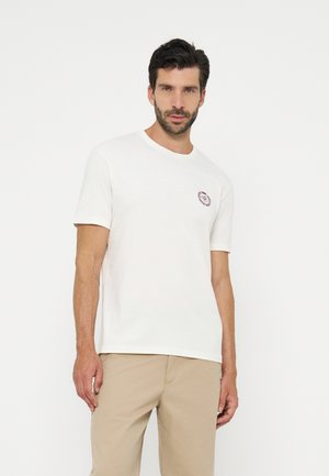 Homme portant un t-shirt blanc uni à manches courtes avec un petit logo sur la poitrine et un pantalon beige, debout sur un fond clair.