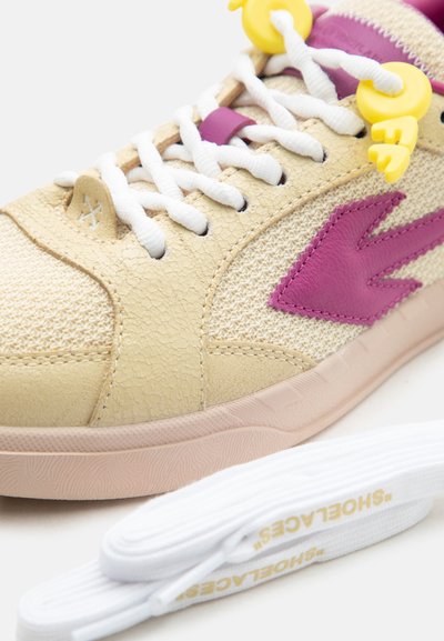 Μπέζ και μοβ αθλητικό παπούτσι με λευκά κορδόνια, κίτρινες διακοσμητικές χάντρες κορδονιών, υφασμένη σόλα και τυλιγμένα λευκά ανταλλακτικά κορδόνια με χρυσό λογότυπο κοντά.