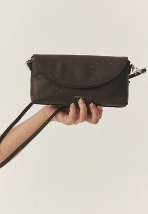 Roka tur tumši brūnu ādas somiņu ar atloku aizdari un plānu regulējamu siksnu uz neitrālas krāsas fona.