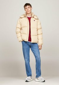 Beige puffer jacket with a zip closure and hood, paired with a red shirt and blue jeans. Model wears gray and white sneakers.
