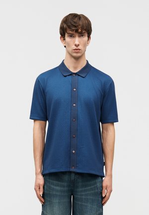 Jeune homme immobile portant une chemise bleue texturée à manches courtes avec col et un jean bleu délavé.