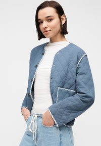 Νεαρή γυναίκα που φοράει ένα μπλε καπιτονέ μπουφάν πάνω από λευκή μπλούζα, με τα χέρια στις τσέπες ανοιχτό μπλε τζιν παντελονιών, σε μονόχρωμο φόντο.