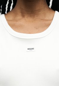 Κοντινό πλάνο του λαιμού και του άνω μέρος του στήθους ενός ατόμου που φοράει λευκό ριμπ τοπ με μια μικρή ετικέτα "Moschino Jeans" στο λαιμό.