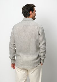 Camicia grigia chiaro a maniche lunghe con un tessuto testurizzato, polsini con bottoni e colletto classico, mostrata da dietro. Infilerata in pantaloni chiari.