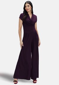 Purple jumpsuit with a wrap-style bodice, cap sleeves, wide legs, and pleated detailing. Worn with burgundy high-heeled sandals.