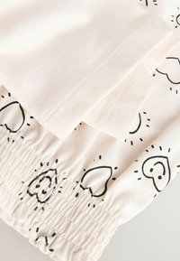 Fehér pamutszövet fekete szív- és napmintákkal. Ráncolt elasztikus derékrész, látható varrási részletekkel. Simogató tapintású felület.