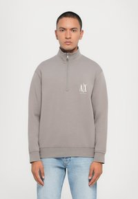 Sudadera de felpa gris con cuello alto, cierre de media cremallera y un logo blanco en el pecho. Textura suave y corte relajado.