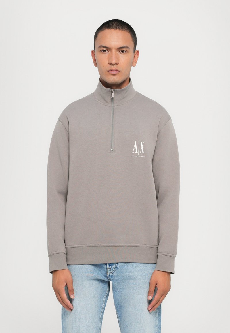 Sudadera de felpa gris con cuello alto, cierre de media cremallera y un logo blanco en el pecho. Textura suave y corte relajado.