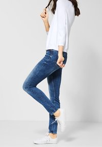 Úzké tmavě modré džínové kalhoty s vybledlým designem, doplněné bílými teniskami a světlě šedým svetrem. Viditelný detail nastavitelných pasových oček.