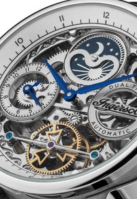 Montre squelette avec un boîtier en acier inoxydable argenté, aiguilles bleues, mécanismes d'engrenages complexes et un affichage des phases de lune avec des motifs d'étoiles.