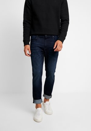 Mand iført mørkeblå oprullede jeans, hvide sneakers og en sort langærmet skjorte, der står mod en ensfarvet baggrund.