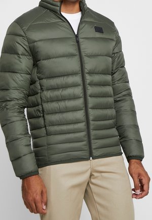 Hombre con chaqueta acolchada con cremallera y capucha de color verde oliva, camisa blanca debajo y pantalones beige sobre un fondo liso.