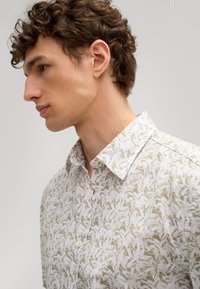 Leichtes, kurzärmeliges Hemd mit einem weißen Blumenmuster auf beigem Hintergrund. Verfügt über einen Button-Down-Kragen und Ein-Knopf-Verschlüsse.