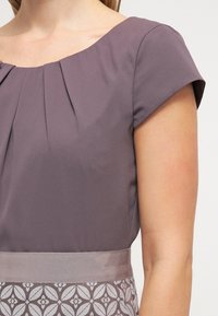 Grijze korte jurk met een geplooide halslijn en een gemusterde tailleband. Glad materiaal met een subtiele glans. Bloemmotief in lichtere grijze accenten.