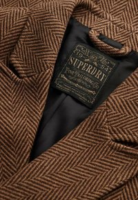 Blazer marrón de espiga con un acabado texturizado, que cuenta con un forro interior negro y una etiqueta de logo que dice "Superdry Fine Tailoring Co."