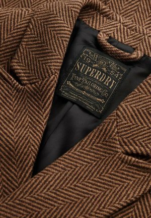 Blazer marrón de espiga con un acabado texturizado, que cuenta con un forro interior negro y una etiqueta de logo que dice "Superdry Fine Tailoring Co."