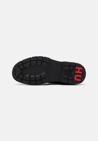 Semelle de chaussure noire avec des rainures profondes et un marquage rouge "HU" sur le talon, texte noir "HUGO" sur l’orteil, sur fond blanc.