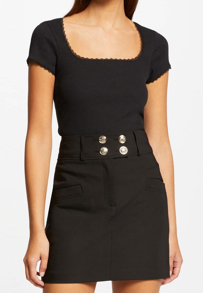 Femme portant un haut noir côtelé à manches courtes avec bordure en dentelle et une jupe noire taille haute avec quatre boutons dorés décoratifs.